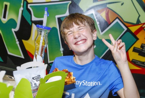 Boy grinning eating pizza from his birthday party box and making a peace sign with his fingers.