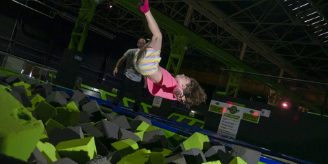 Girl suspended mid air upside down as she is about to fall into a pit of green and black foam squares in indoor play area.